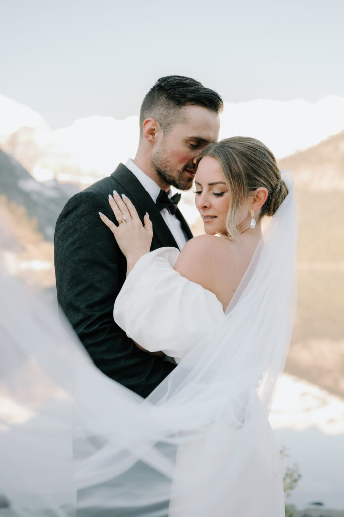 bride-and-groom-embracing-with-flowing-veil-styled-lake-louise-wedding-mountain-backdrop-banff-wedding-photography